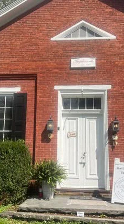 Sunny exterior of a historic red-brick storefront: white paneled door with transom, black shutters and lantern sconces, potted plants on a stone step and a sidewalk sandwich-board sign.