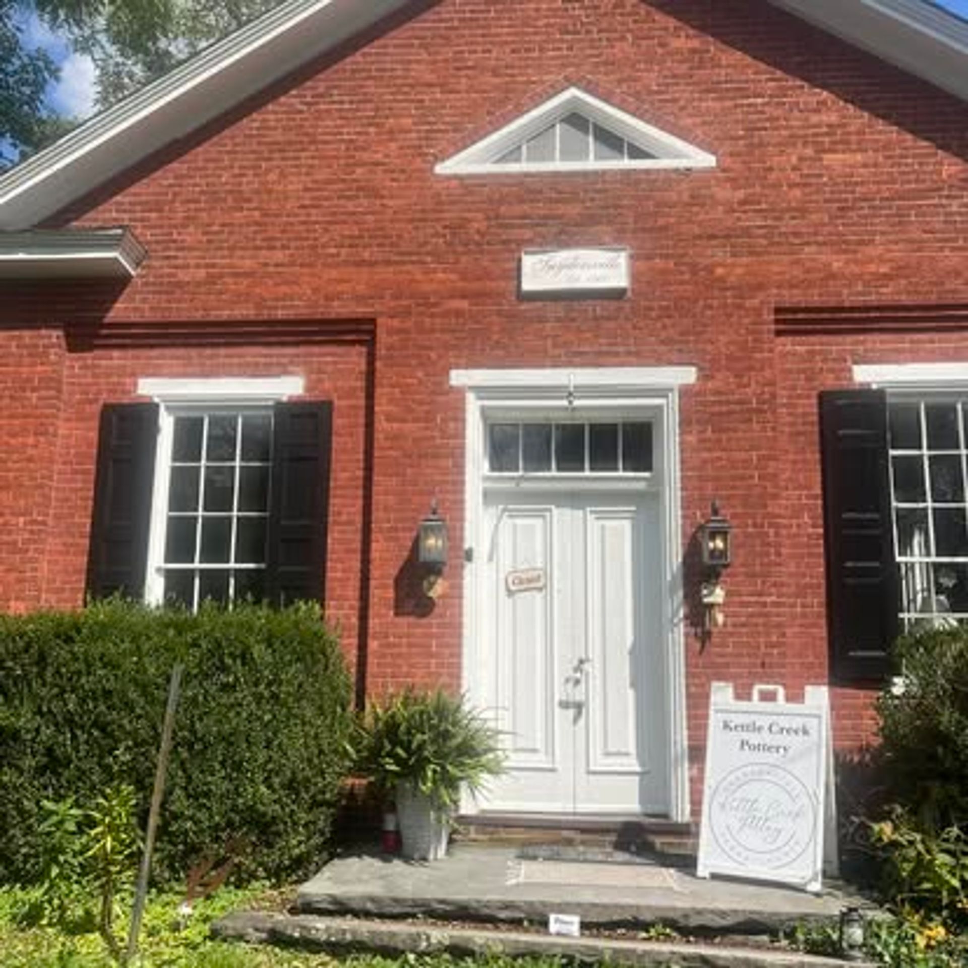 Sunny exterior of a historic red-brick storefront: white paneled door with transom, black shutters and lantern sconces, potted plants on a stone step and a sidewalk sandwich-board sign.