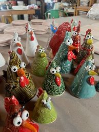 Playful group of colorful glazed handmade ceramic rooster and chicken figurines with wide eyes and textured details arranged on a pottery studio worktable.