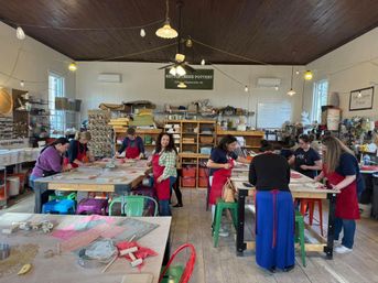 Busy community pottery studio with people in aprons shaping clay at long worktables, shelves of glazes and tools, colorful stools and hanging lights under a dark wood ceiling.