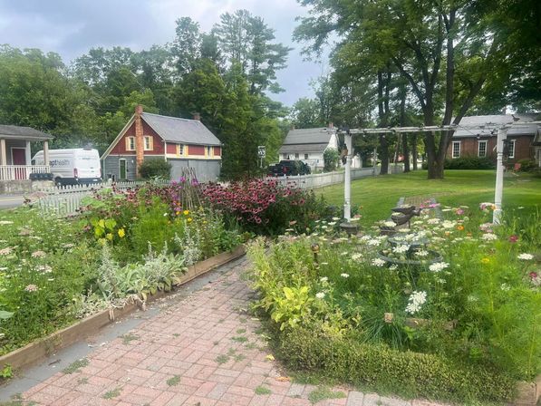 Front-yard wildflower garden and brick path leading past a white picket fence to a tree-lined small-town street with historic houses.