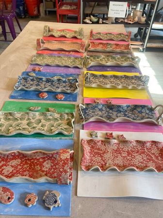 Rows of colorful handmade wavy-edged ceramic trays with floral and geometric patterns set on bright paper backings on a pottery studio worktable, with small matching ceramic tiles nearby.
