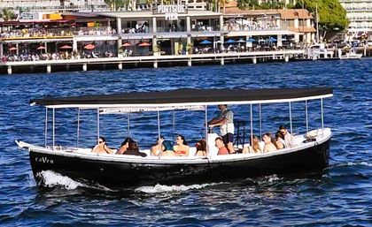 Covered black tour boat carrying passengers across blue harbor waters past a bustling waterfront pier with outdoor dining.