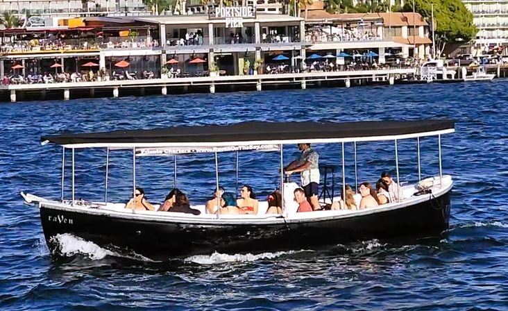 Covered black tour boat carrying passengers across blue harbor waters past a bustling waterfront pier with outdoor dining.