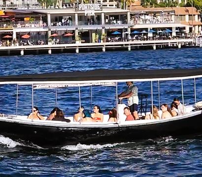 Covered black tour boat carrying passengers across blue harbor waters past a bustling waterfront pier with outdoor dining.