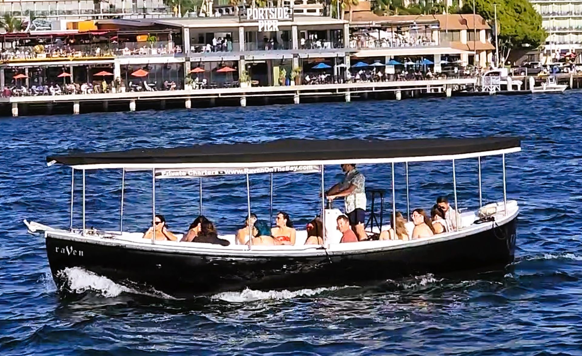 Covered black tour boat carrying passengers across blue harbor waters past a bustling waterfront pier with outdoor dining.