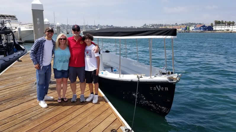 Four people smiling on a wooden marina dock next to a covered small boat in a sunny coastal harbor with sailboats and shoreline in the background
