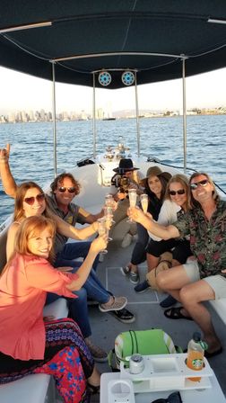 Group of friends toasting with champagne on a small canopy boat in a harbor at sunset, with an urban skyline across the water.