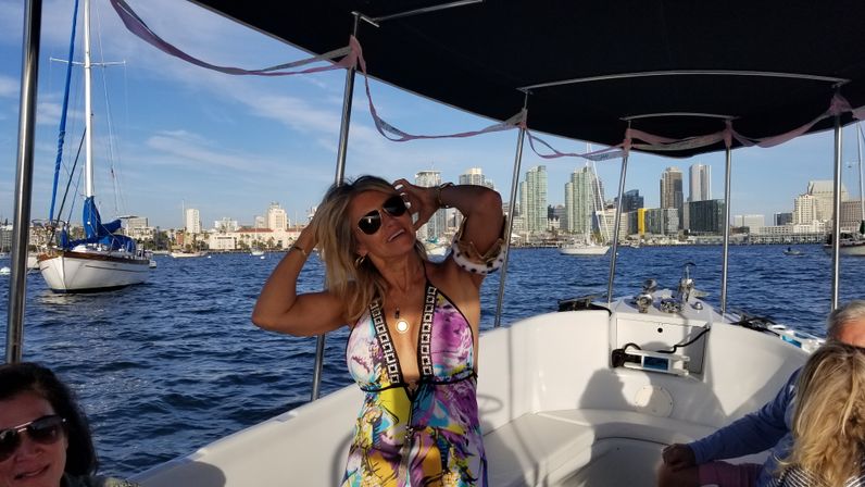 Smiling woman in a colorful sundress and sunglasses enjoying a sunny boat ride with sailboats and a coastal city skyline in the harbor