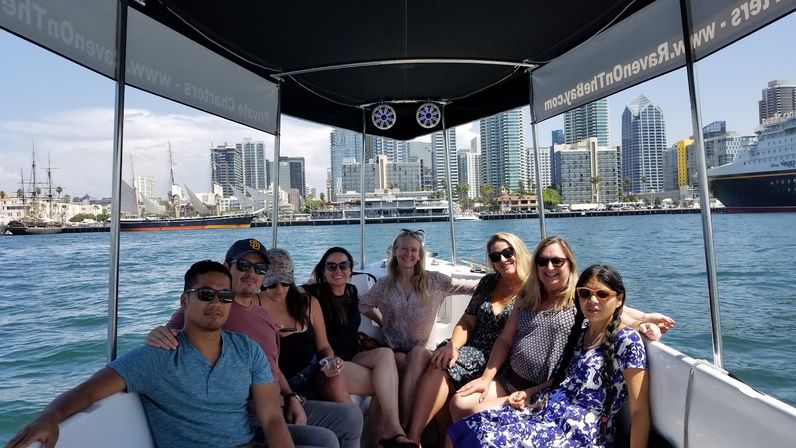 Smiling group of people relaxing on a shaded boat tour in sunny San Diego Harbor with the downtown skyline, tall buildings, and historic ships in the background.