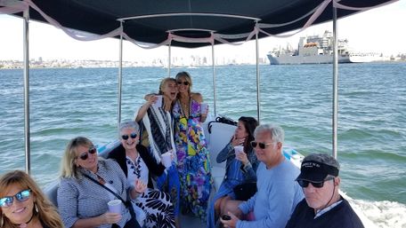 Friends on a covered party boat holding drinks and smiling, cruising a sunny harbor with a city skyline and large ship on the horizon.