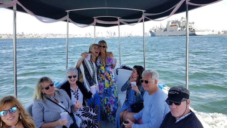 Friends on a covered party boat holding drinks and smiling, cruising a sunny harbor with a city skyline and large ship on the horizon.
