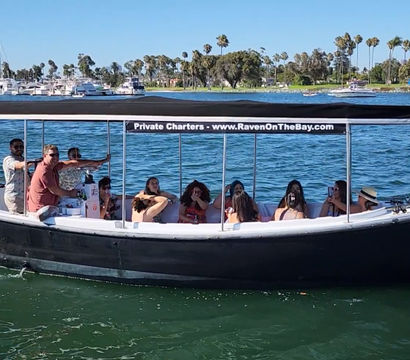 Black covered charter boat carrying a group on a sunny, palm-lined bay with sailboats and a marina in the background.
