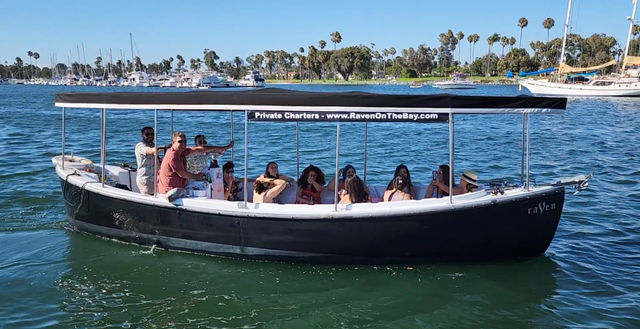 Black covered charter boat carrying a group on a sunny, palm-lined bay with sailboats and a marina in the background.