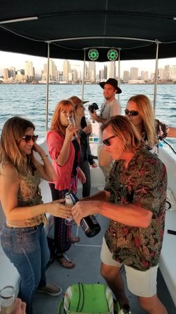 Group of friends sipping champagne and toasting on a covered party boat with a downtown skyline and harbor at sunset.