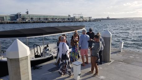 Group of adults boarding a covered boat at a sunny marina pier, concrete dock pilings and industrial waterfront with a large ship on the distant horizon.