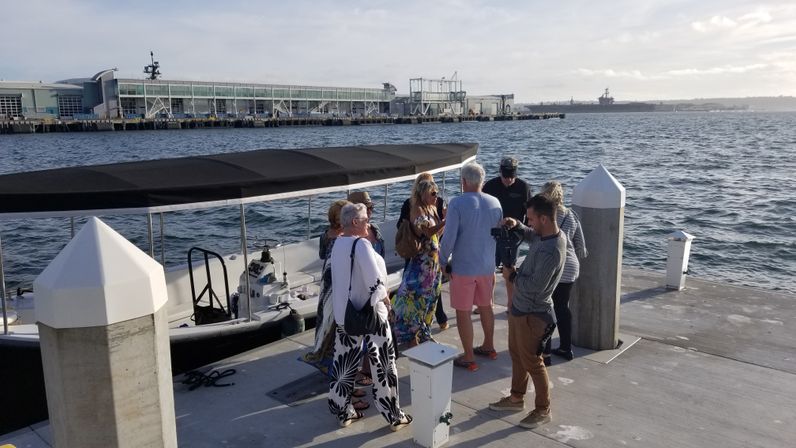 Group of adults boarding a covered boat at a sunny marina pier, concrete dock pilings and industrial waterfront with a large ship on the distant horizon.