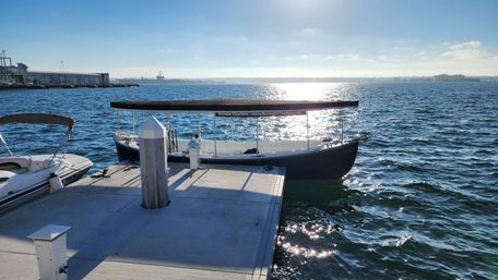 Sunlit covered passenger boat moored at a concrete dock in a sparkling blue bay, marina pier and distant harbor silhouetted under a clear sky