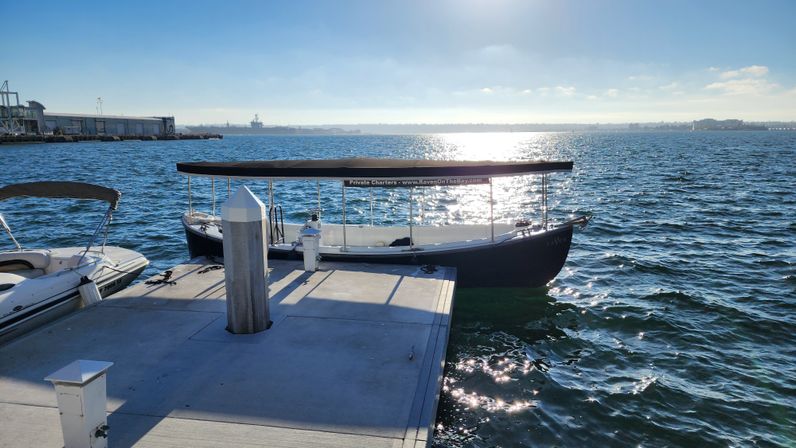 Sunlit covered passenger boat moored at a concrete dock in a sparkling blue bay, marina pier and distant harbor silhouetted under a clear sky