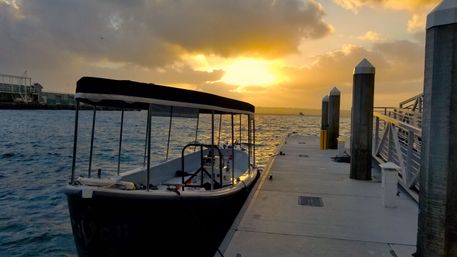Covered boat moored at a concrete dock in a harbor as a golden sunset breaks through clouds, reflecting on rippling water and wooden pilings.