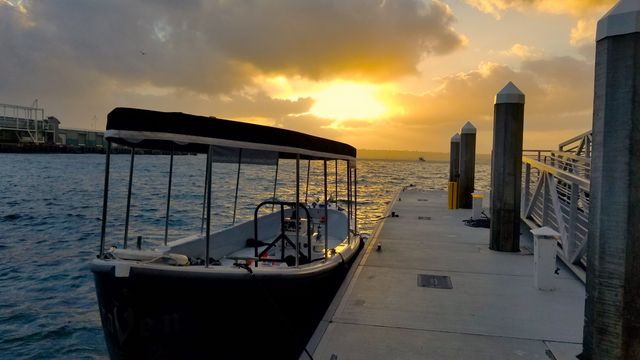 Covered boat moored at a concrete dock in a harbor as a golden sunset breaks through clouds, reflecting on rippling water and wooden pilings.