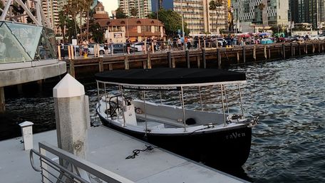 Small black-and-white covered tour boat moored at a bustling urban waterfront marina, with a wooden pier, palm trees, and high-rise condos lining the lively promenade at golden hour.