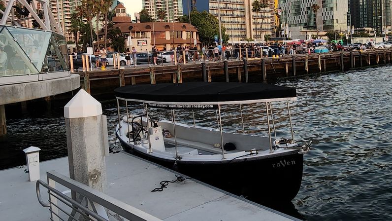 Small black-and-white covered tour boat moored at a bustling urban waterfront marina, with a wooden pier, palm trees, and high-rise condos lining the lively promenade at golden hour.
