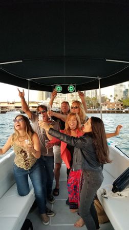 Friends cheering and toasting with champagne on a covered party boat near a waterfront city skyline and palm trees during a harbor cruise.