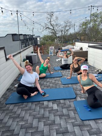 Five women doing rooftop yoga on blue mats on an urban rooftop terrace with string lights, outdoor lounge seating, and city buildings beyond.