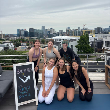 Six women smiling on a rooftop yoga/pilates mat after a bachelorette workout with the Nashville skyline in the background.