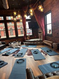 Cozy vintage‑chic urban loft yoga studio with blue mats and Pilates rings on wood floor, exposed brick walls, large arched windows, red velvet curtains and globe pendant lights.
