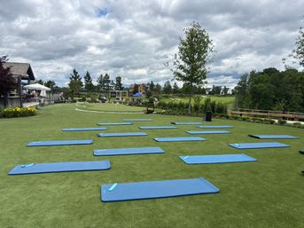 Blue yoga mats arranged on green turf for an outdoor fitness class beside a clubhouse and putting green at a golf course, cloudy sky and homes in the distance