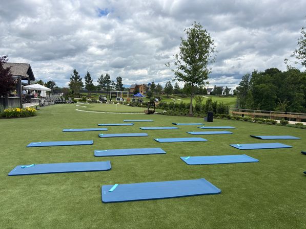 Blue yoga mats arranged on green turf for an outdoor fitness class beside a clubhouse and putting green at a golf course, cloudy sky and homes in the distance