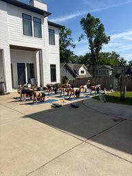 Outdoor group yoga class on blue mats performing leg lifts on a sunny suburban driveway beside a modern white house and tall trees.