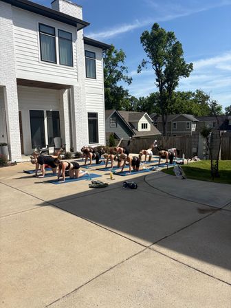 Outdoor group yoga class on blue mats performing leg lifts on a sunny suburban driveway beside a modern white house and tall trees.