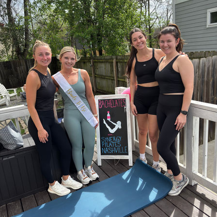 Four women in athletic wear pose on a wooden deck beside a yoga mat and chalkboard sign advertising a bachelorette Pilates class in Nashville; one woman wears a bride-to-be sash.