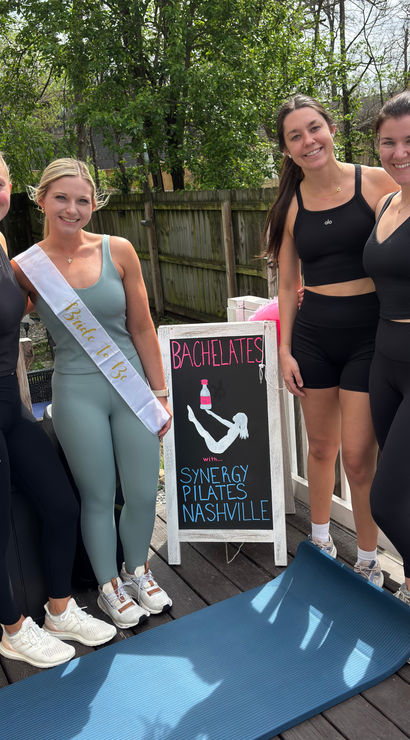 Four women in athletic wear pose on a wooden deck beside a yoga mat and chalkboard sign advertising a bachelorette Pilates class in Nashville; one woman wears a bride-to-be sash.