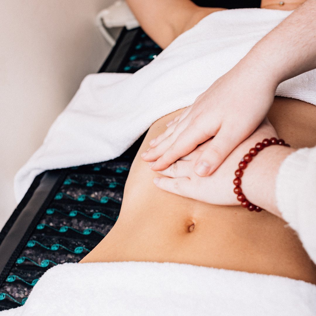 Close-up of spa abdominal massage on a treatment table – hands pressing the lower belly over white towels, red bead bracelet visible, wellness therapy scene