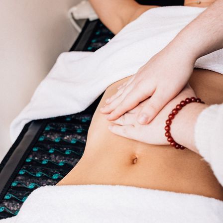 Close-up of spa abdominal massage on a treatment table – hands pressing the lower belly over white towels, red bead bracelet visible, wellness therapy scene