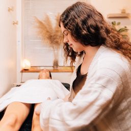 Relaxing spa scene with a practitioner massaging a client’s legs on a treatment table in a cozy wellness room with warm lighting, salt lamp, and pampas grass.
