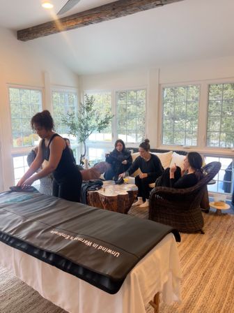 Bright sunroom-style living room with large windows and exposed beam; a wellness practitioner readies a heated blanket on a massage table while four women and a dog relax on wicker chairs around a rustic wood coffee table with forest views outside.