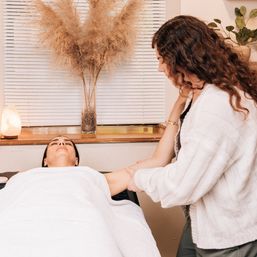 Client relaxed on a spa table receiving an arm massage from a therapist in a cozy treatment room with white linens, a salt lamp and dried pampas grass.