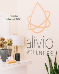Bright minimalist wellness studio reception nook with a pale-peach abstract droplet logo on a cream wall, white console with a gold-trim table lamp, potted succulents, black candle, and a tall snake plant.
