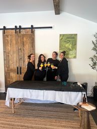 Four women in black toasting mimosas beside a covered massage table in a bright wellness studio with a rustic sliding barn door and green abstract art