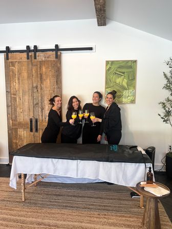 Four women in black toasting mimosas beside a covered massage table in a bright wellness studio with a rustic sliding barn door and green abstract art