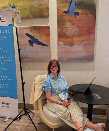 Conference attendee with glasses and badge seated at a round table with a laptop in a hotel conference area, colorful bird wall art panels and a promotional banner in the background.