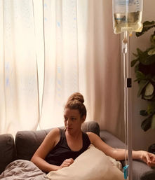 Person reclining on a gray sofa in a sunlit living room, receiving an IV infusion from a hanging saline bag with curtains and a potted plant in the background.