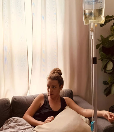 Person reclining on a gray sofa in a sunlit living room, receiving an IV infusion from a hanging saline bag with curtains and a potted plant in the background.