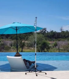 Sunlit poolside scene with a person reclining on a white lounge chair in shallow water under a bright turquoise umbrella, clear blue pool, a wheeled pole nearby, and green trees beyond the pool deck.