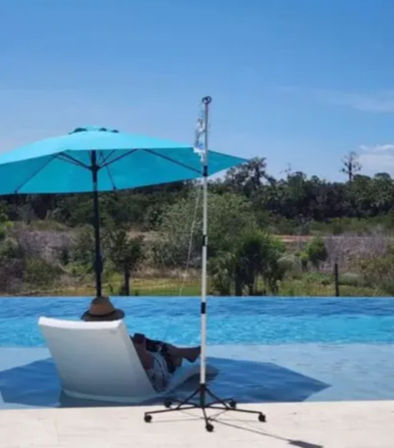 Sunlit poolside scene with a person reclining on a white lounge chair in shallow water under a bright turquoise umbrella, clear blue pool, a wheeled pole nearby, and green trees beyond the pool deck.